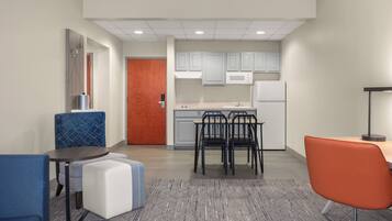 A modern kitchen with white cabinets, a refrigerator, and a microwave. There are two dining tables with chairs, one with a blue chair and the other with an orange chair. A small white ottoman is placed between the tables.