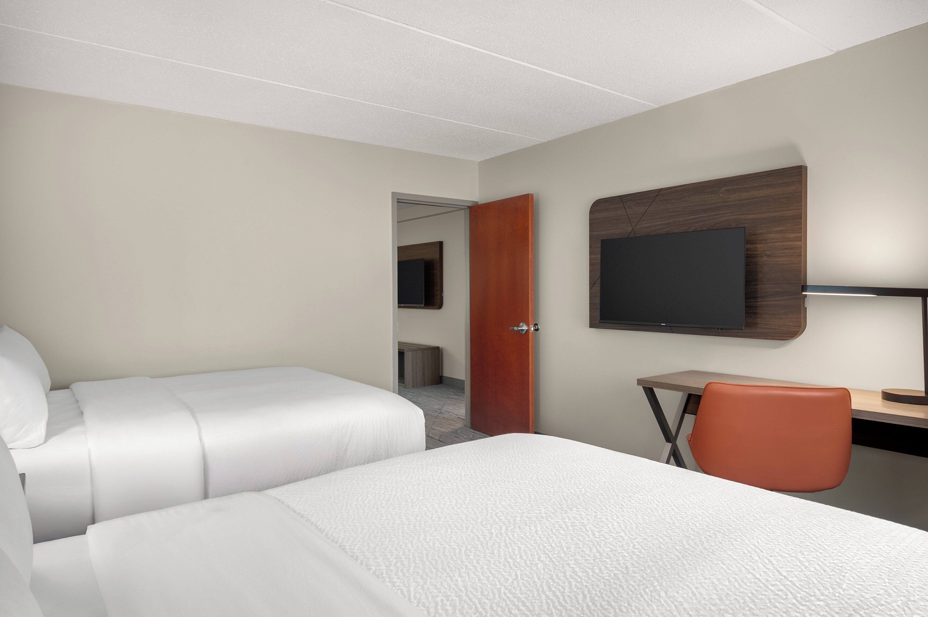Habitación de hotel con dos camas, un escritorio con silla, un televisor y una puerta que da a otra habitación.