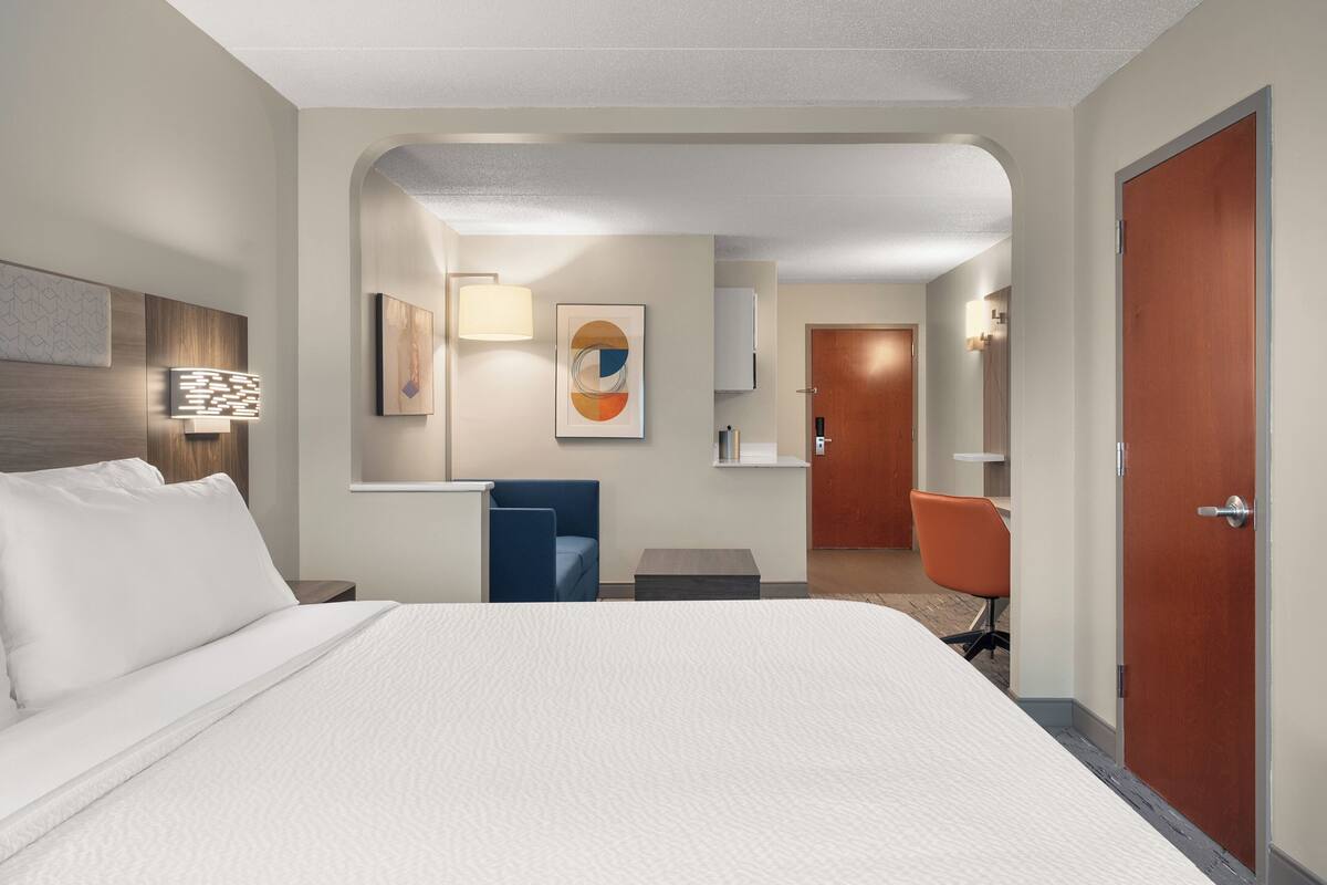 A hotel room with a large bed, a desk, a chair, a wardrobe, and a bathroom visible through an archway.