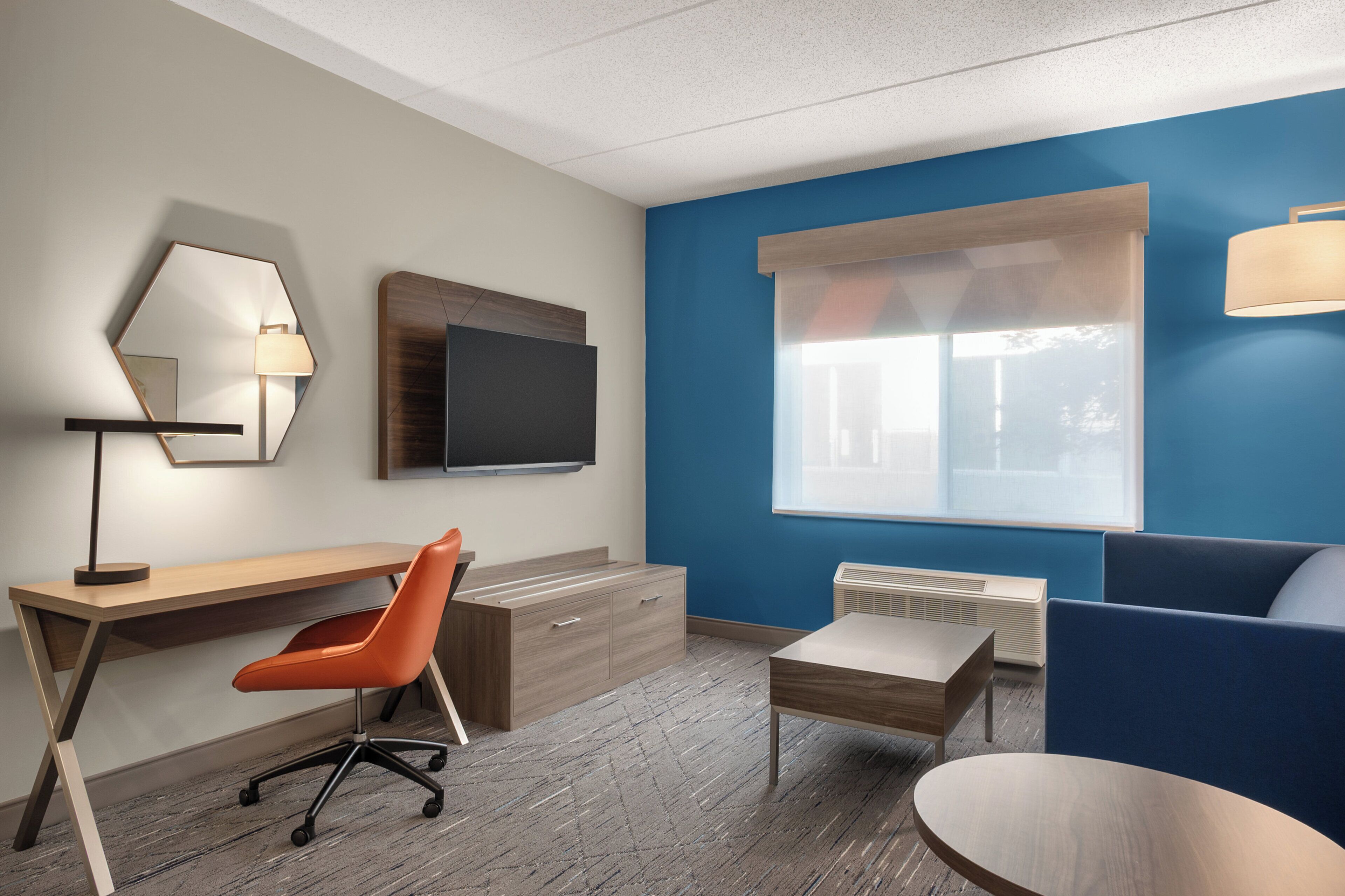 Una habitación de hotel moderna con un escritorio de madera, una silla de oficina naranja, un televisor de pantalla plana, una pared de acento azul, un ventanal con persianas, una pequeña mesa y un sofá azul.