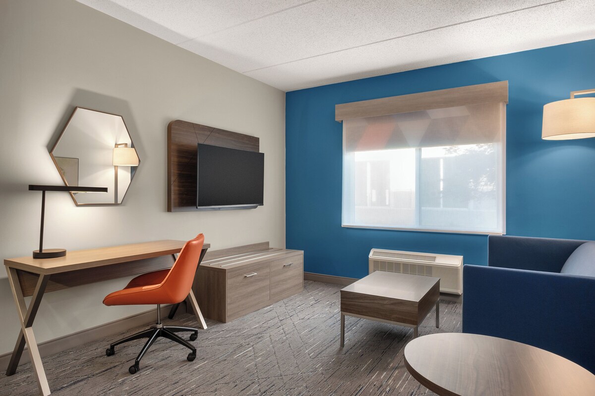 Quarto de hotel moderno com escrivaninha de madeira, cadeira de escritório laranja, TV de tela plana, parede de destaque azul, ampla janela com persianas, pequena mesa e sofá azul.