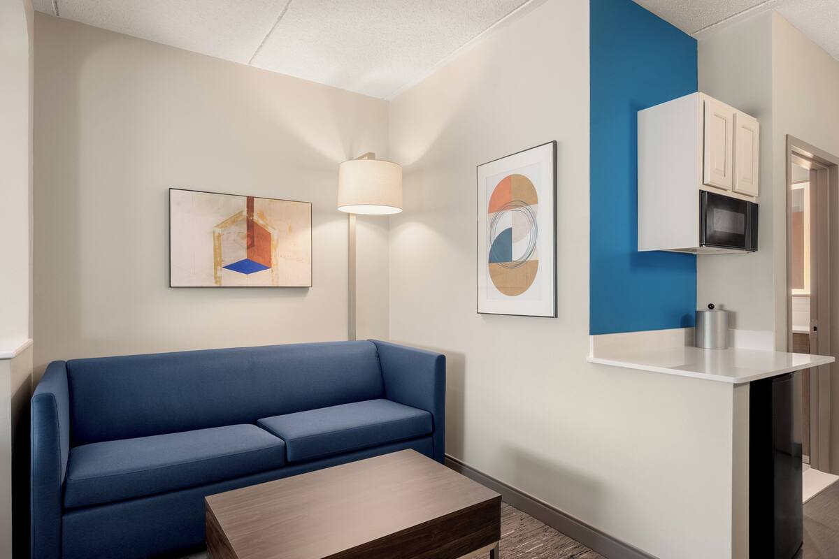 A modern hotel room with a blue sofa, a wooden coffee table, a lamp, and artwork on the walls.