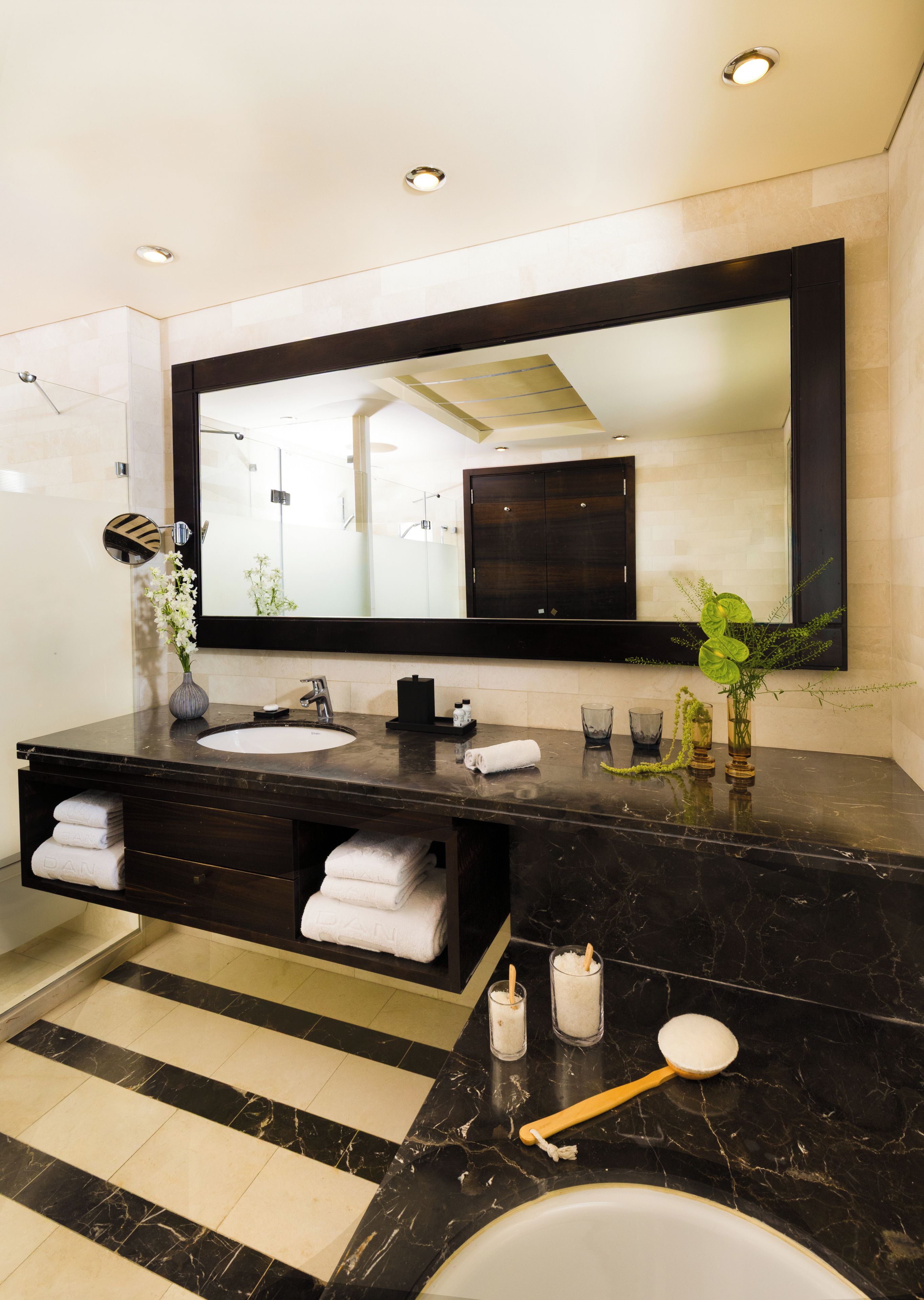 Un baño moderno con un espejo grande, encimera de mármol oscuro, lavabo blanco y mueble de tocador negro.