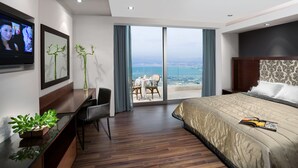 A modern hotel room with a large bed, a desk, a chair, and a view of the city through a glass door. - The Dan Carmel Hotel (Haifa)