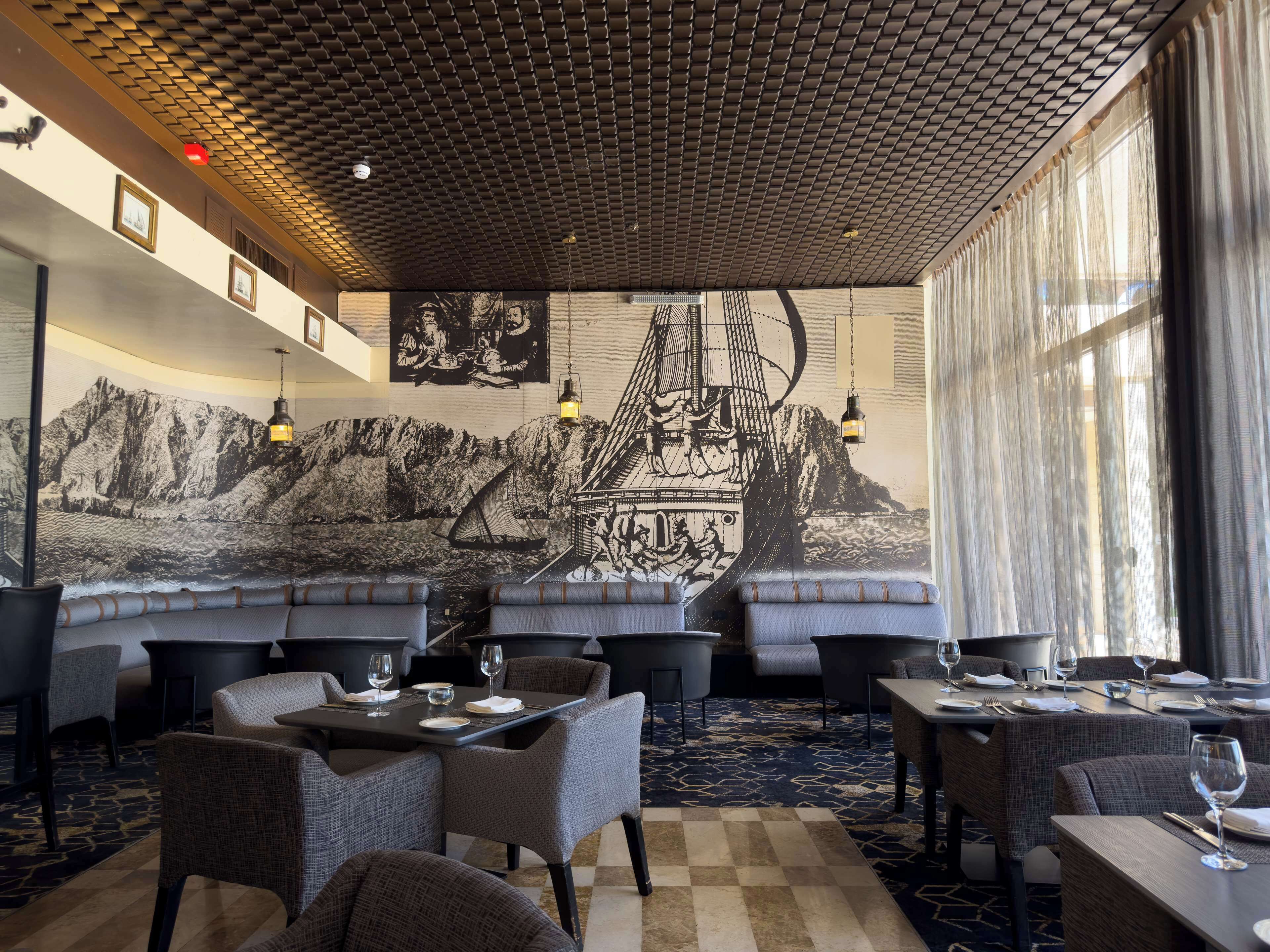 A restaurant with high-backed chairs, tables set for dining, and a large mural depicting a sailing ship on a mountain backdrop.