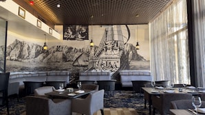 Um restaurante com cadeiras de encosto alto, mesas postas para refeições e um grande mural retratando um navio a vela ao fundo de uma montanha.