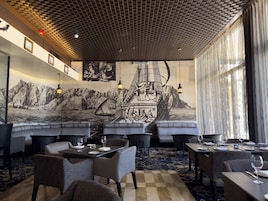 Un restaurante con sillas de respaldo alto, mesas dispuestas para comer y un gran mural que muestra un barco a vela con montañas de fondo.