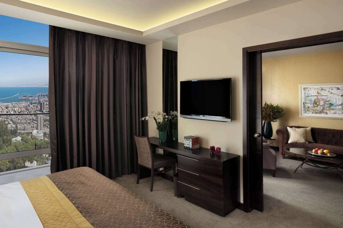 A modern hotel room with a large bed, a desk with a TV, a view of the city, and a cozy sitting area.