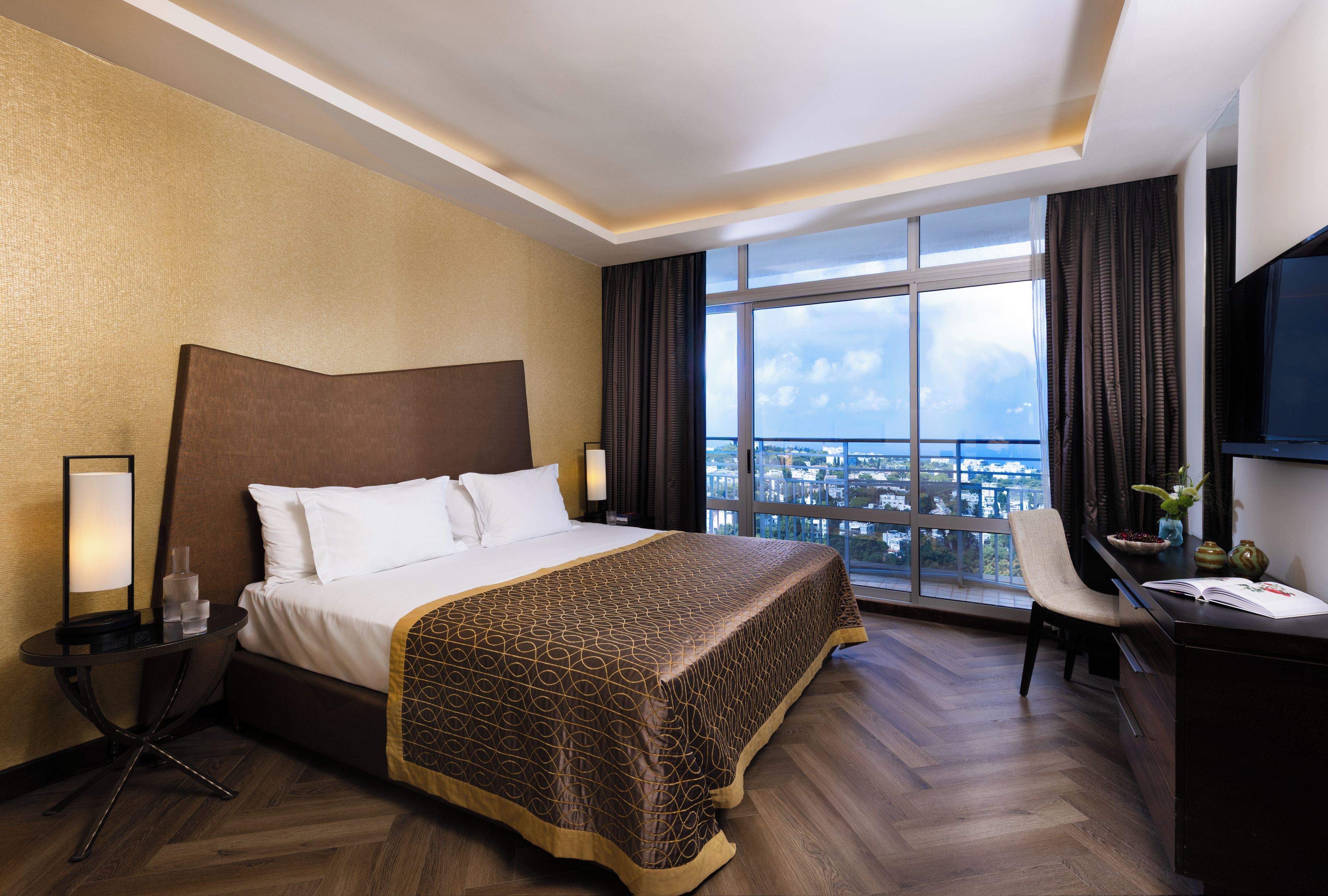 Habitación de hotel con una cama grande, un escritorio con silla, un televisor y vistas a un paisaje urbano a través de un ventanal amplio.