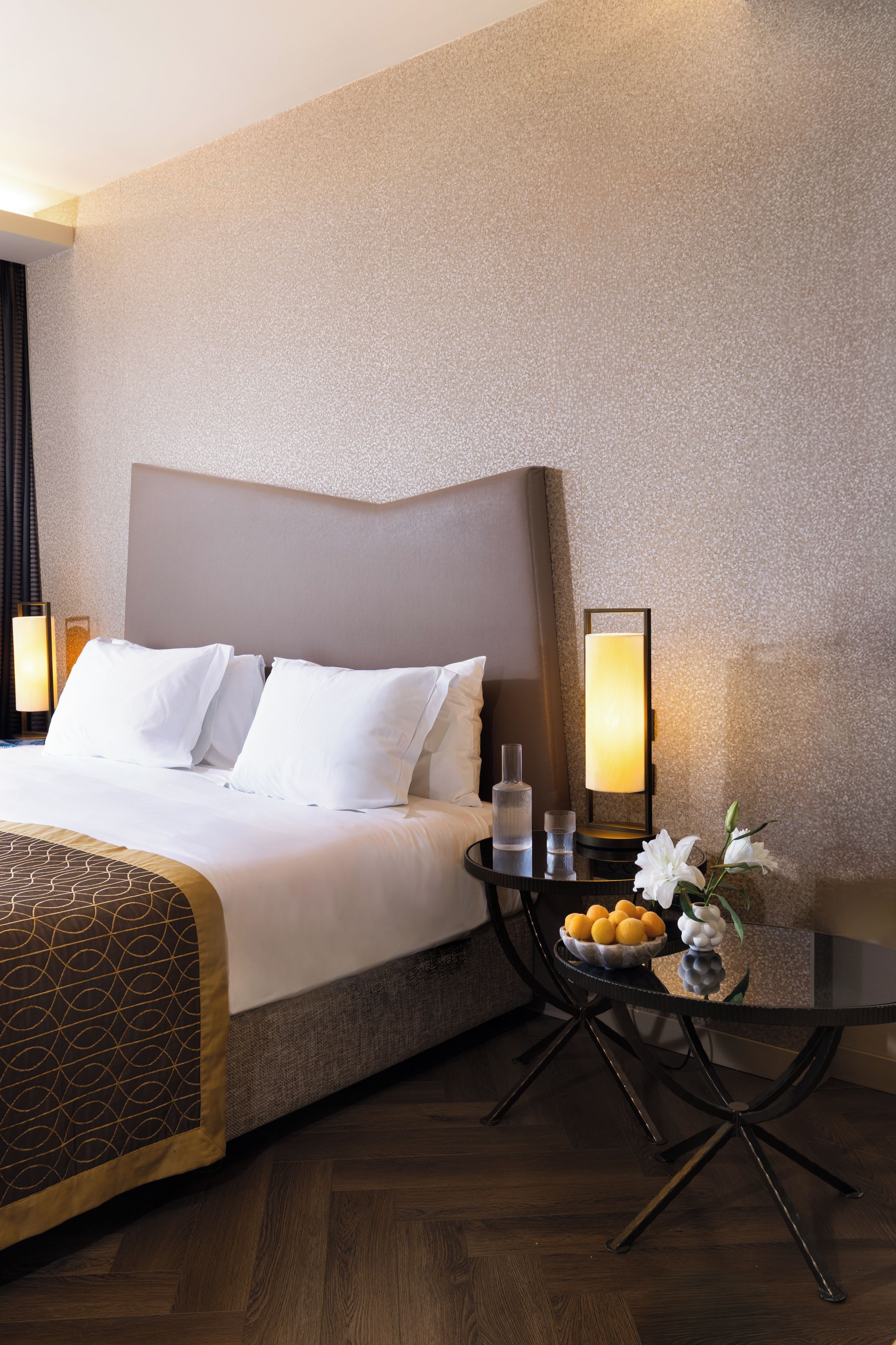 A hotel room with a large bed, bedside lamps, a small table with a vase and fruit, and a wooden floor.
