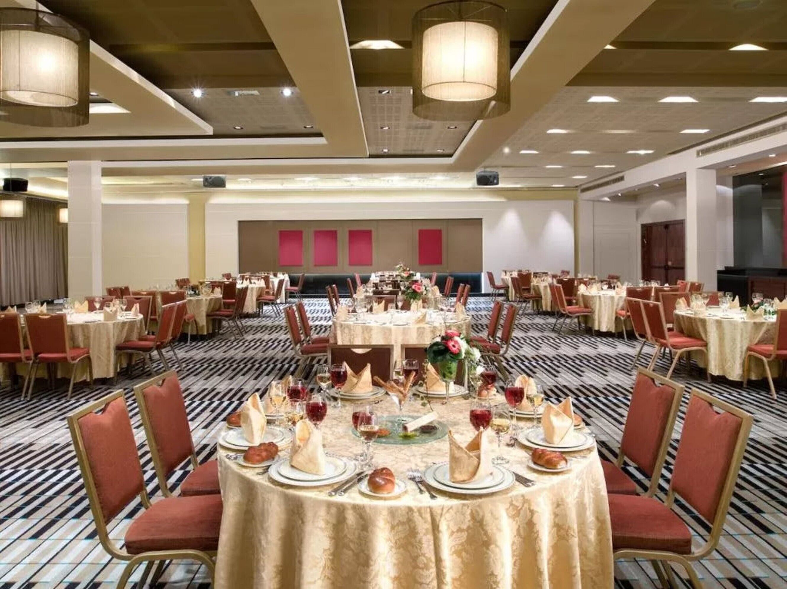 A banquet hall with round tables, red chairs, and a patterned carpet.
