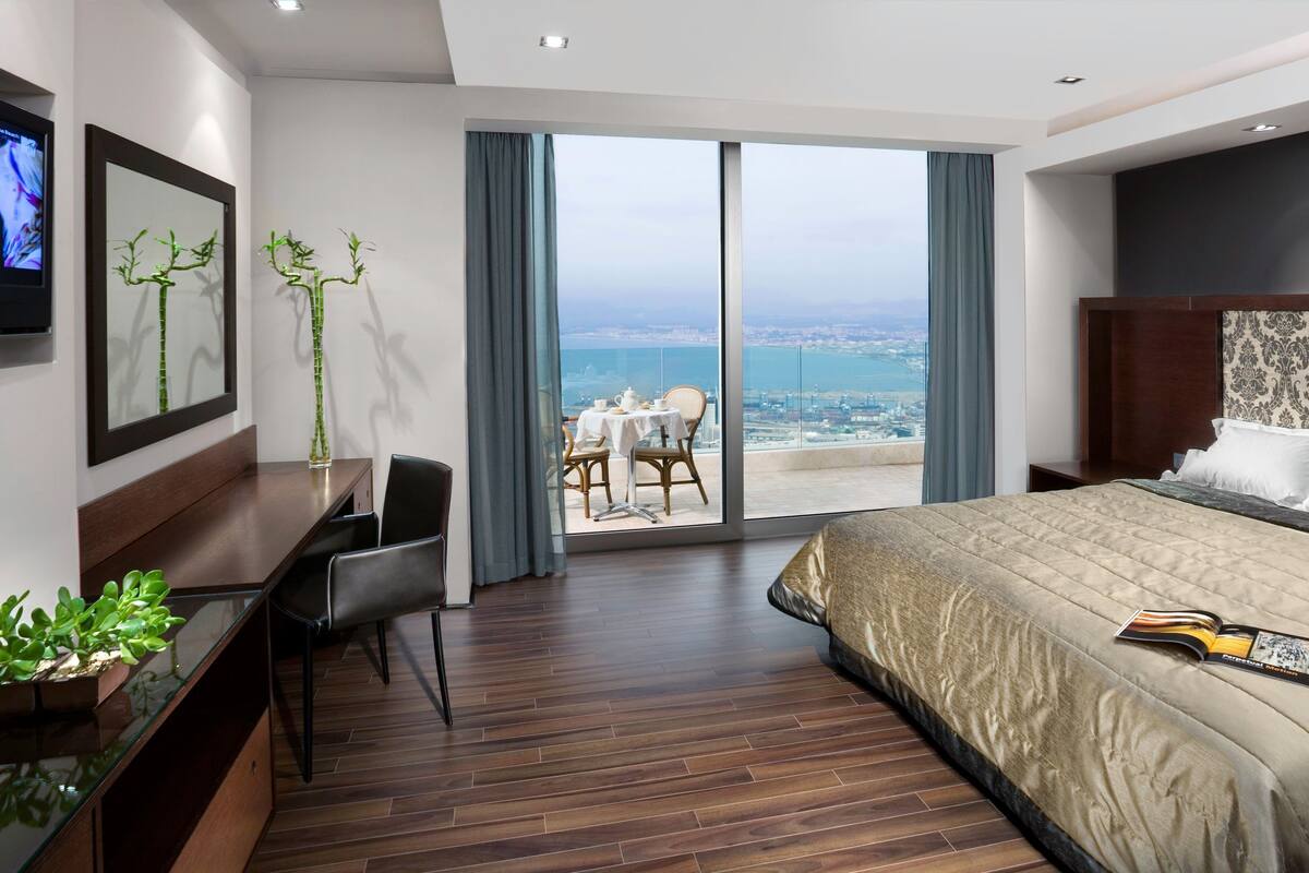 A modern hotel room with a large bed, a desk, a chair, a TV, and a balcony with a city view.