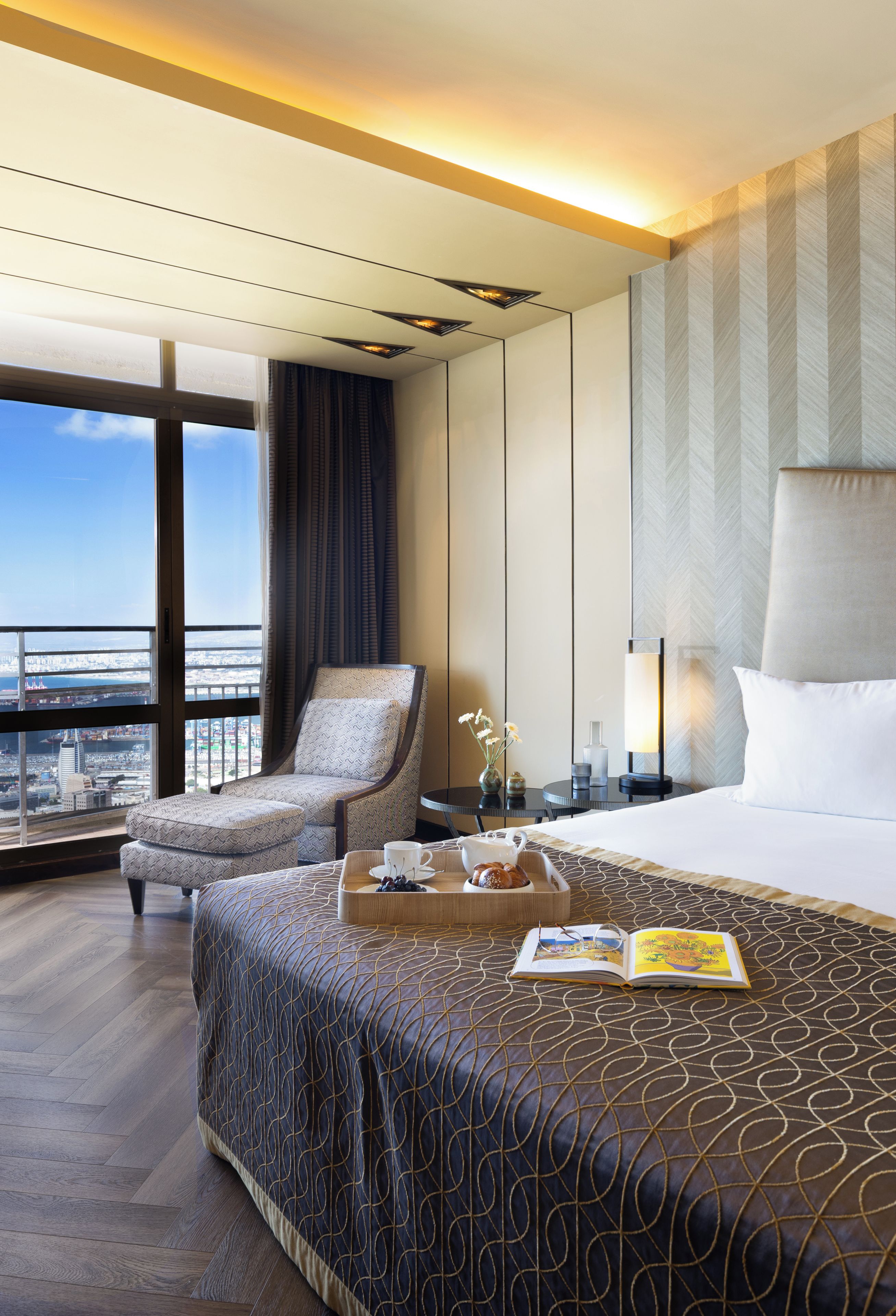 A hotel room with a large bed, a chair, a table with a tray of food, and a view of the city.