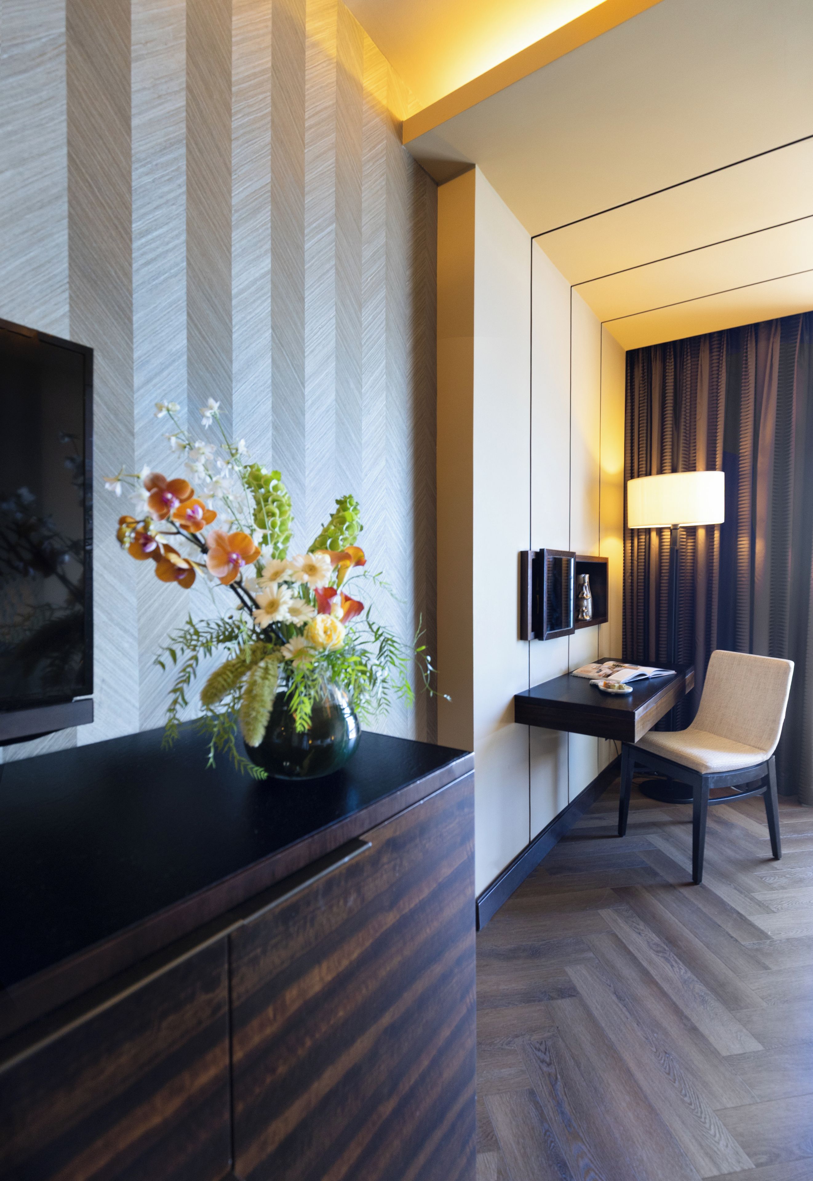A modern hotel room with a wooden floor, a flat-screen TV mounted on the wall, a desk with a lamp, and a chair.