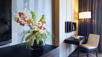 A modern hotel room with a wooden floor, a flat-screen TV mounted on the wall, a desk with a lamp, and a chair.