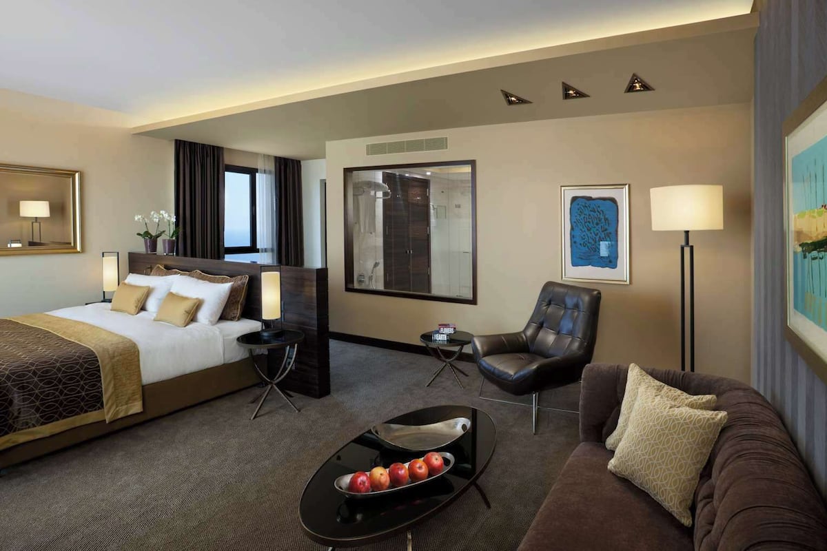 A hotel room with a king-sized bed, a sofa, a chair, a small table with fruit, and artwork on the walls.