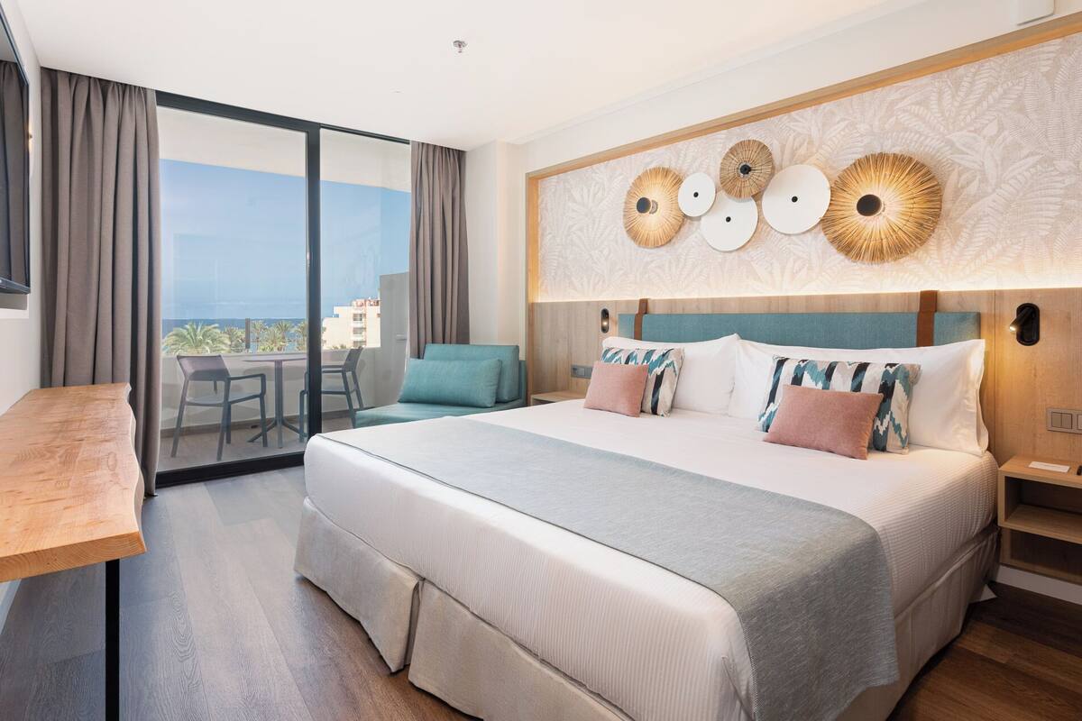 A hotel room with a large bed, a wooden desk, a balcony with a view, and decorative wall art.