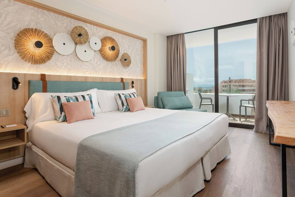 A hotel room with a large bed, a view of the city, and a balcony with a table and chairs.