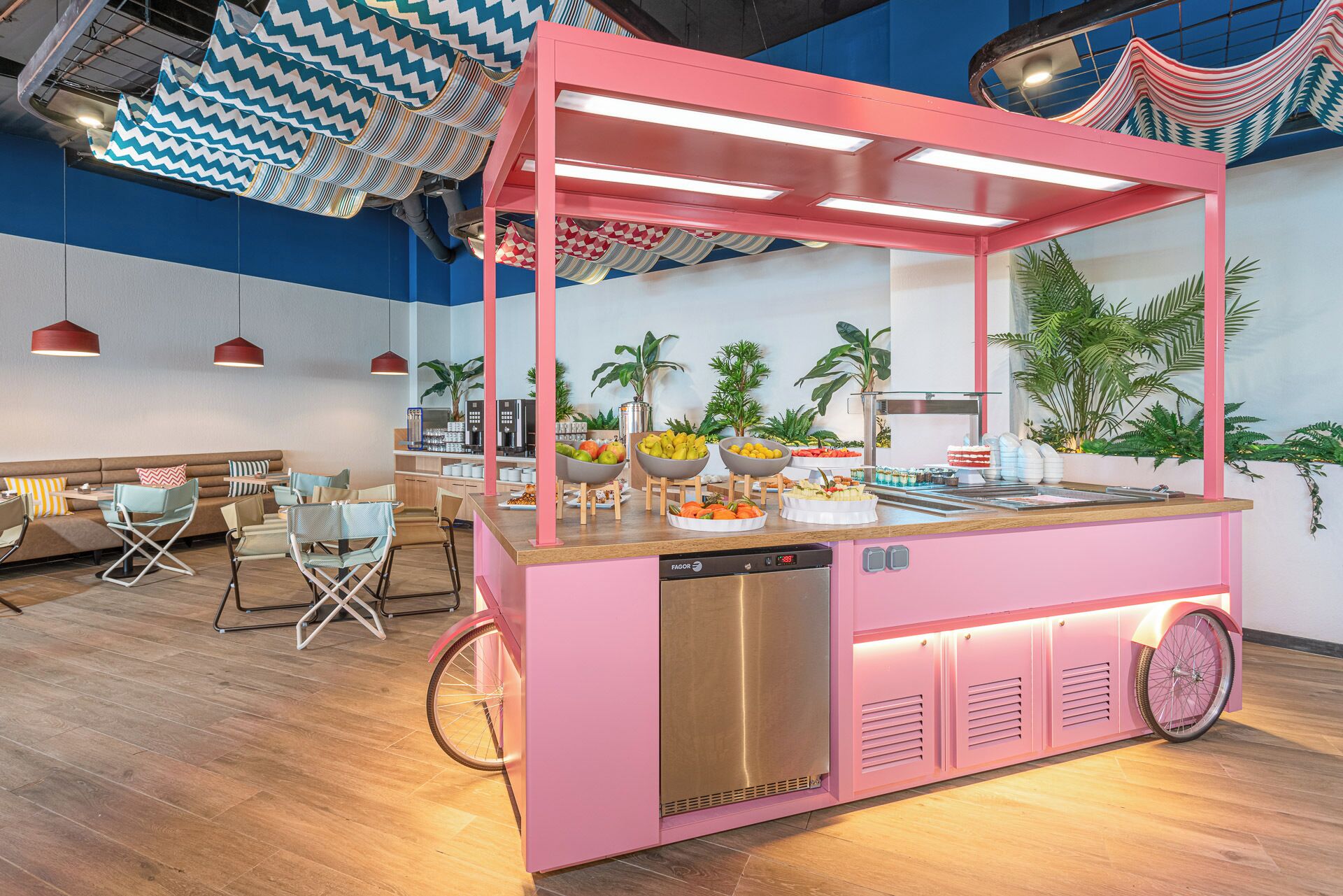 A pink food cart with a stainless steel dishwasher, surrounded by seating and a buffet with fruits and pastries.