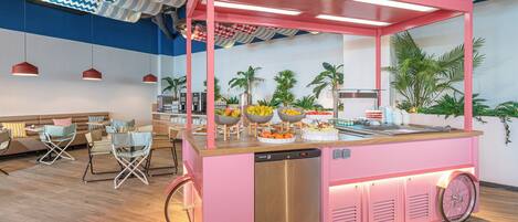 A pink food cart with a stainless steel dishwasher, surrounded by seating and a buffet with fruits and pastries.