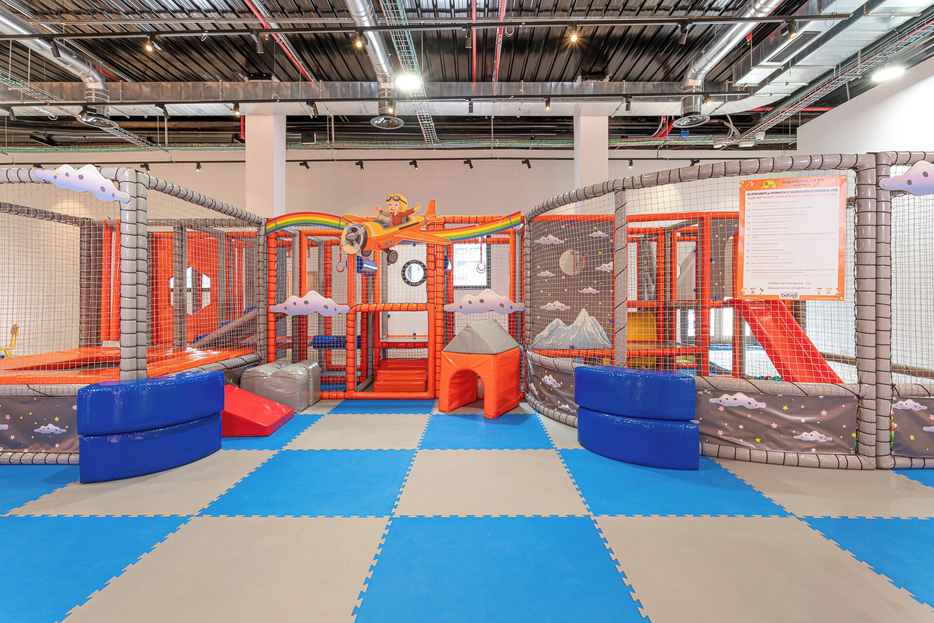Un espace de jeux intérieur doté d’un sol rembourré, de structures de jeux en filet et d’un toboggan central.