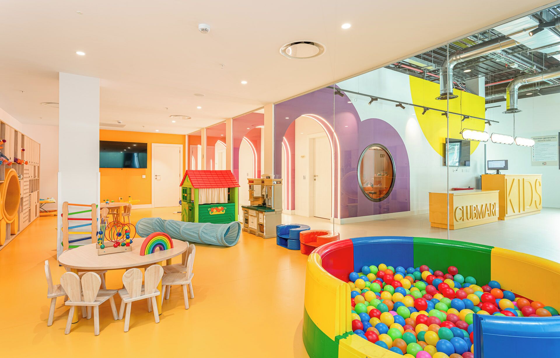 A brightly lit indoor play area with a ball pit, play structures, and a reception desk labeled "KIDS".