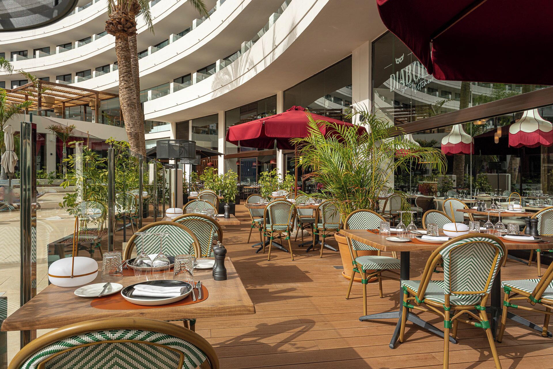 An outdoor dining area with multiple tables and chairs, surrounded by glass walls and red umbrellas.