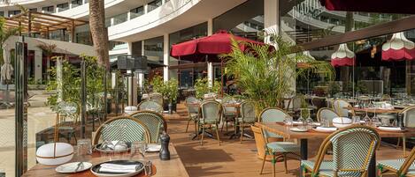 An outdoor dining area with multiple tables and chairs, surrounded by glass walls and red umbrellas.