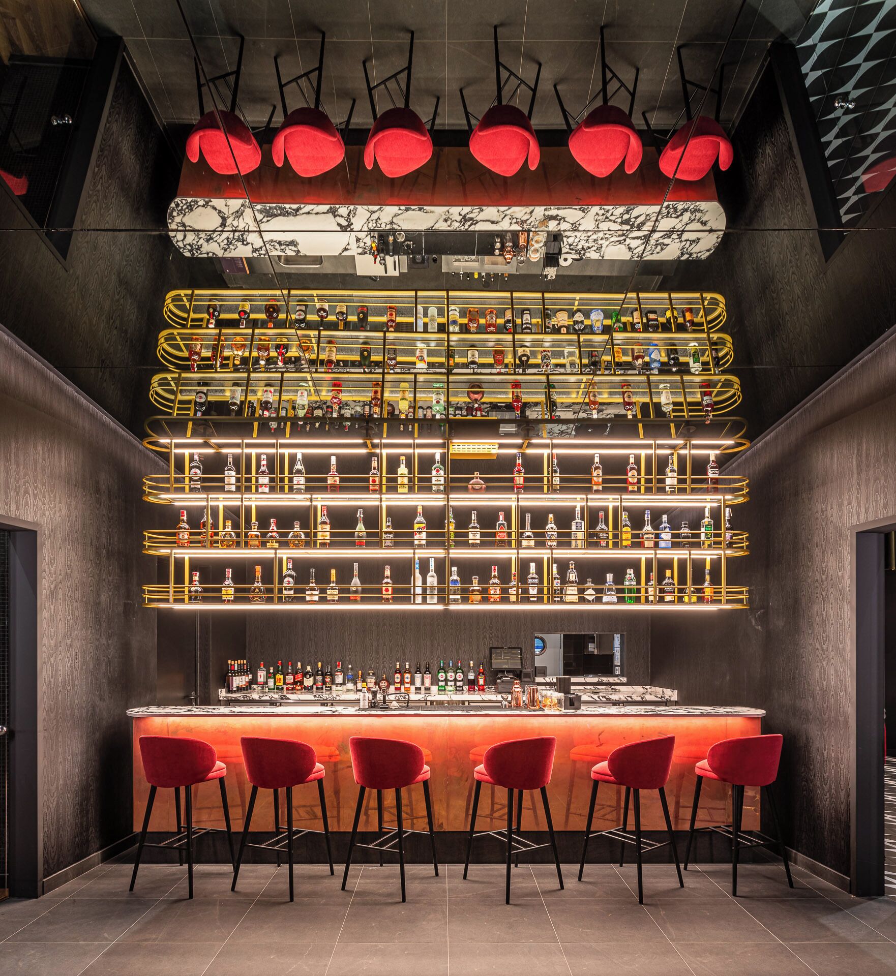 A bar area with multiple shelves of liquor bottles, red bar stools, and a marble countertop.