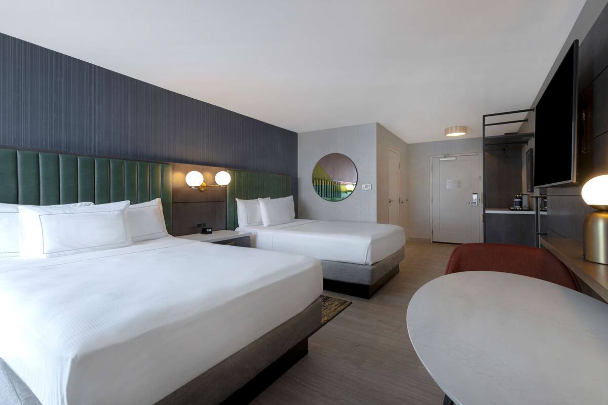 A hotel room with two beds, a round mirror, a TV, and a dining area.