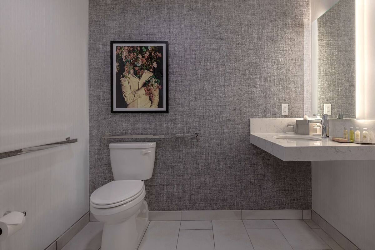 A modern bathroom with a textured gray wall, a framed floral artwork, a white toilet, a white sink with a marble countertop, and a mirror.