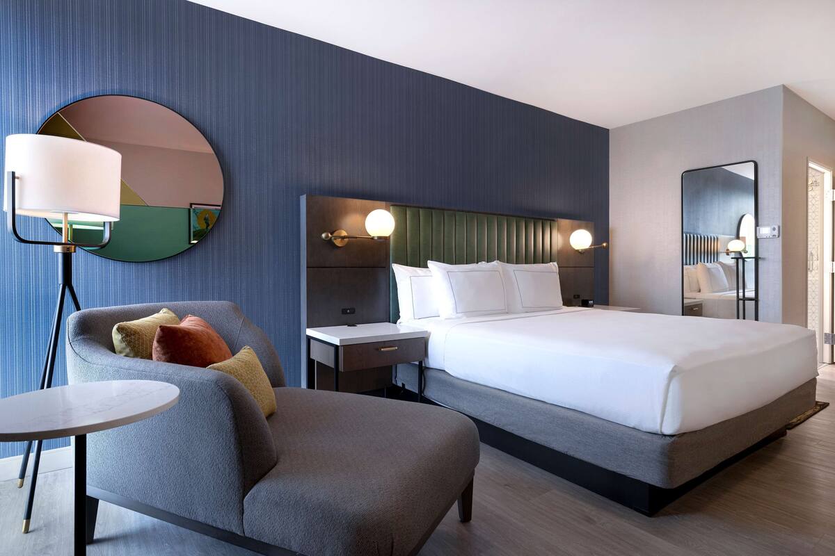 A modern hotel room with a large bed, a round mirror, a desk, and a chair.