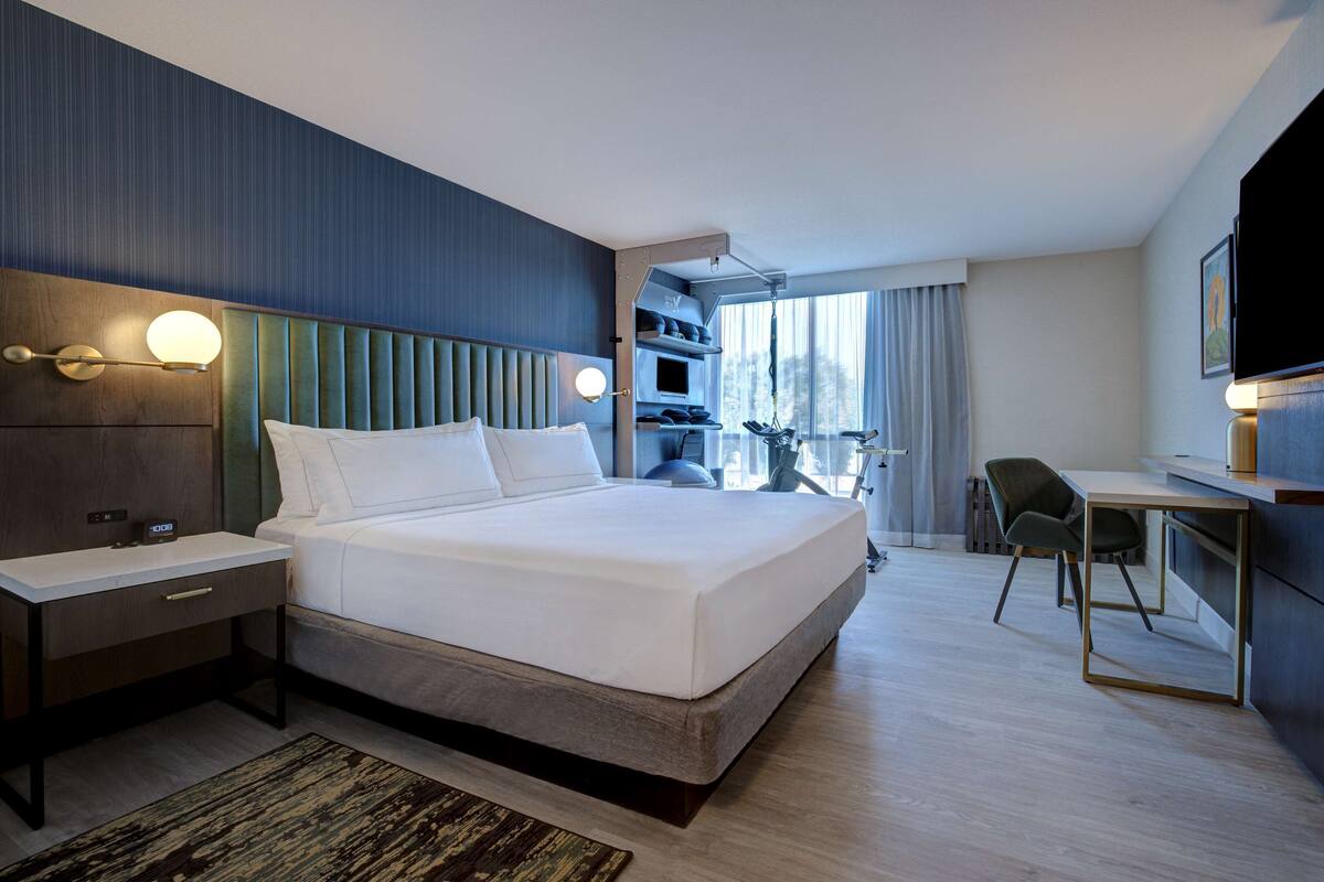 A modern hotel room with a large bed, a desk with a chair, a TV, and a balcony with a view.