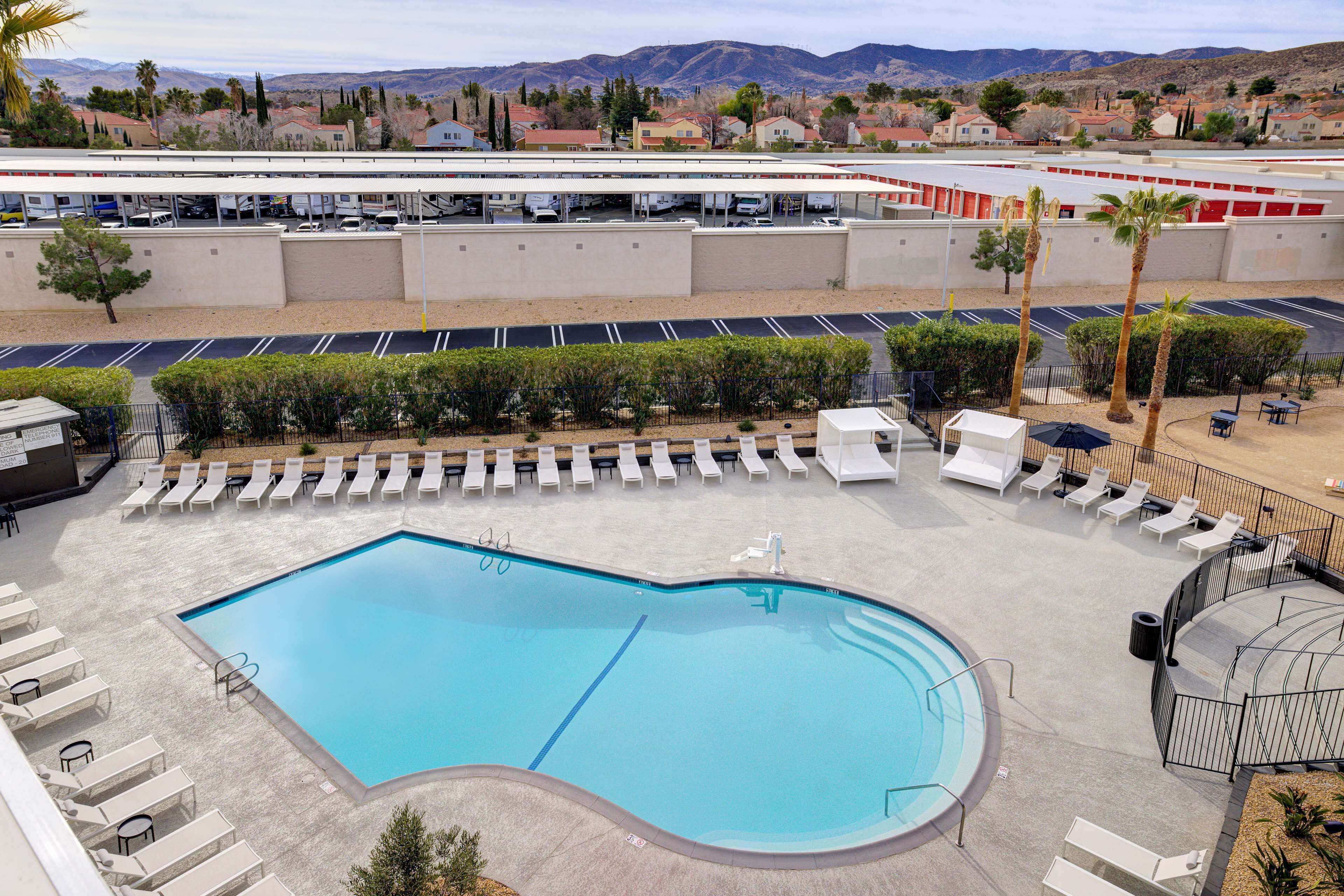 A swimming pool with sun loungers and a parking lot.