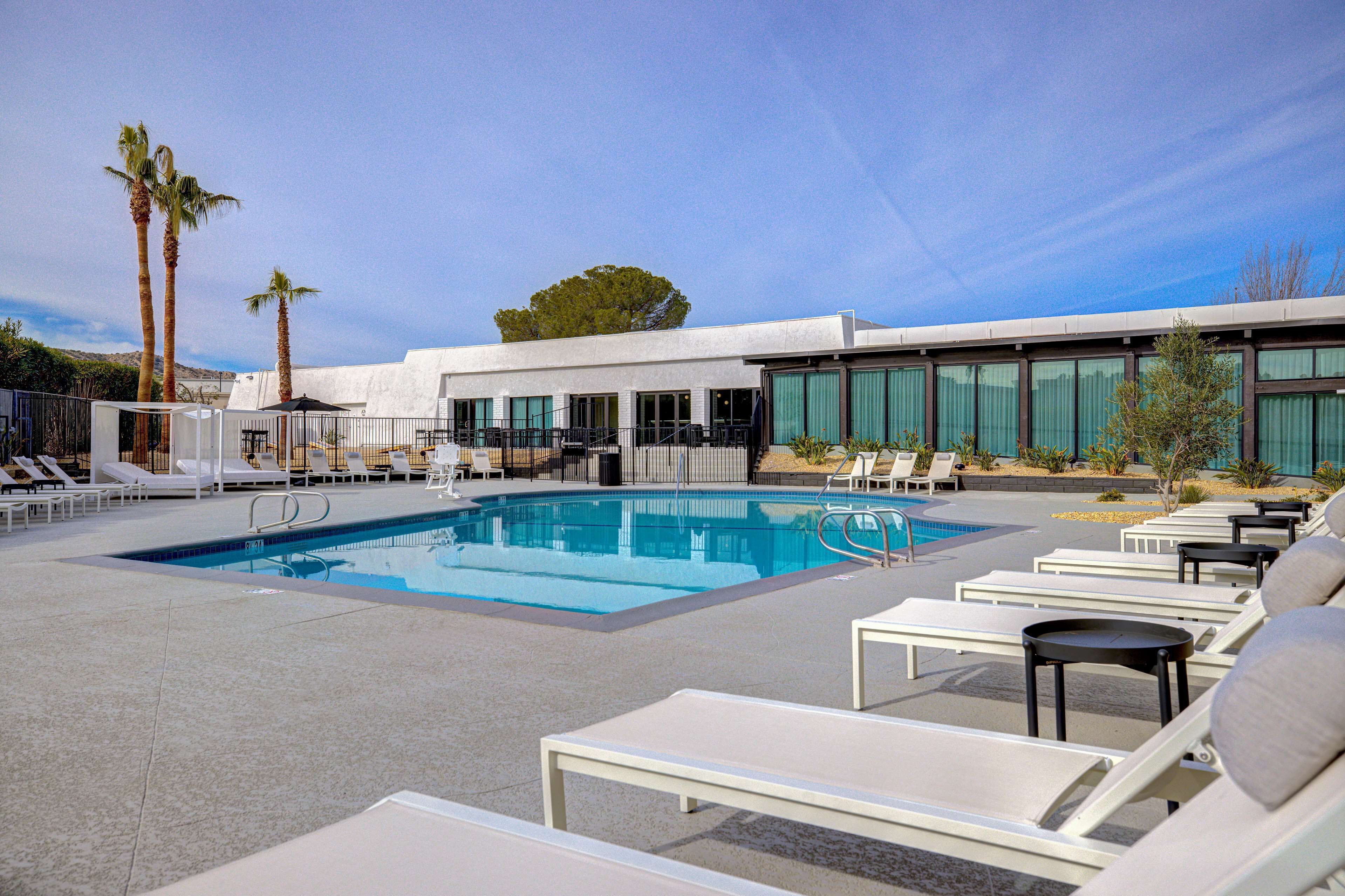 A modern hotel pool area with sun loungers, a swimming pool, and palm trees.