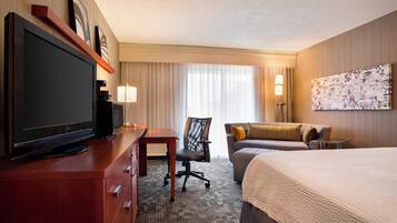 A hotel room with a large flat-screen TV, a desk with a chair, a bed with a large comforter, a sofa with cushions, and a painting on the wall.