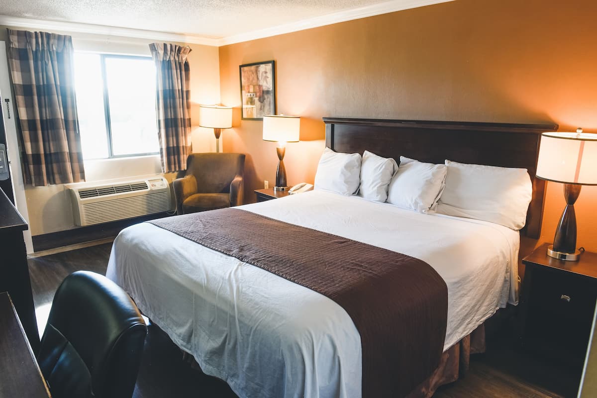 A hotel room with a large bed, two bedside lamps, a desk with a chair, a window with curtains, and a wall-mounted air conditioner.