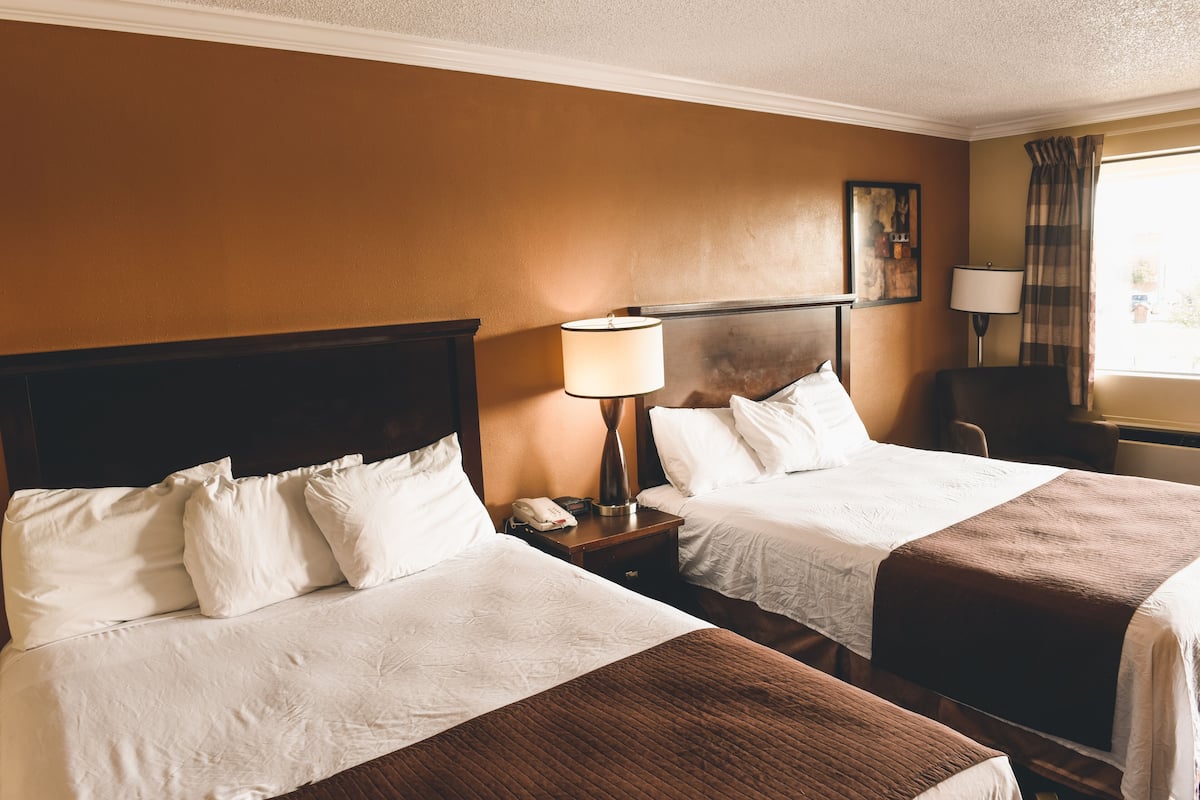 A hotel room with two beds, a nightstand with a lamp, a chair, and a window with curtains.