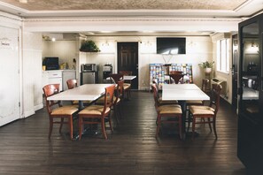 A dining area with a long table and chairs, a kitchen in the background, and a television mounted on the wall.