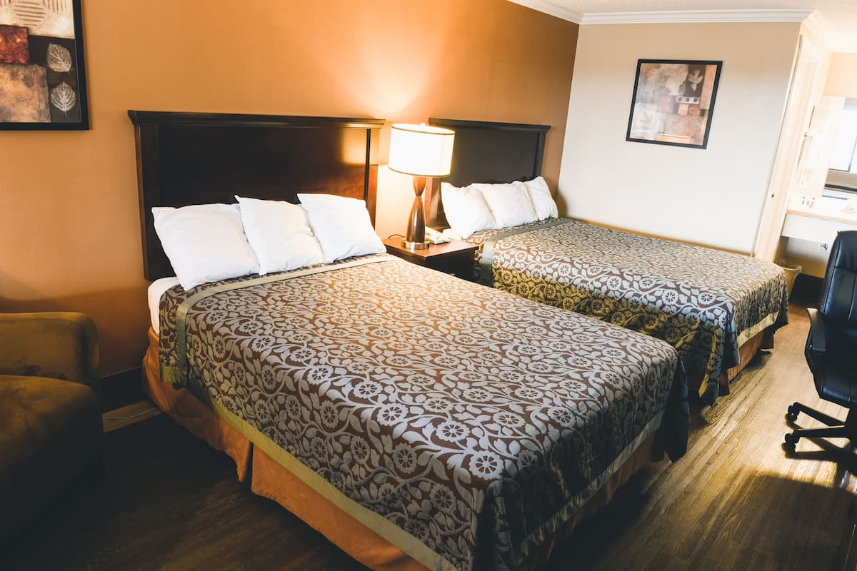 A hotel room with two beds, a nightstand with a lamp, a chair, and a desk with a chair.