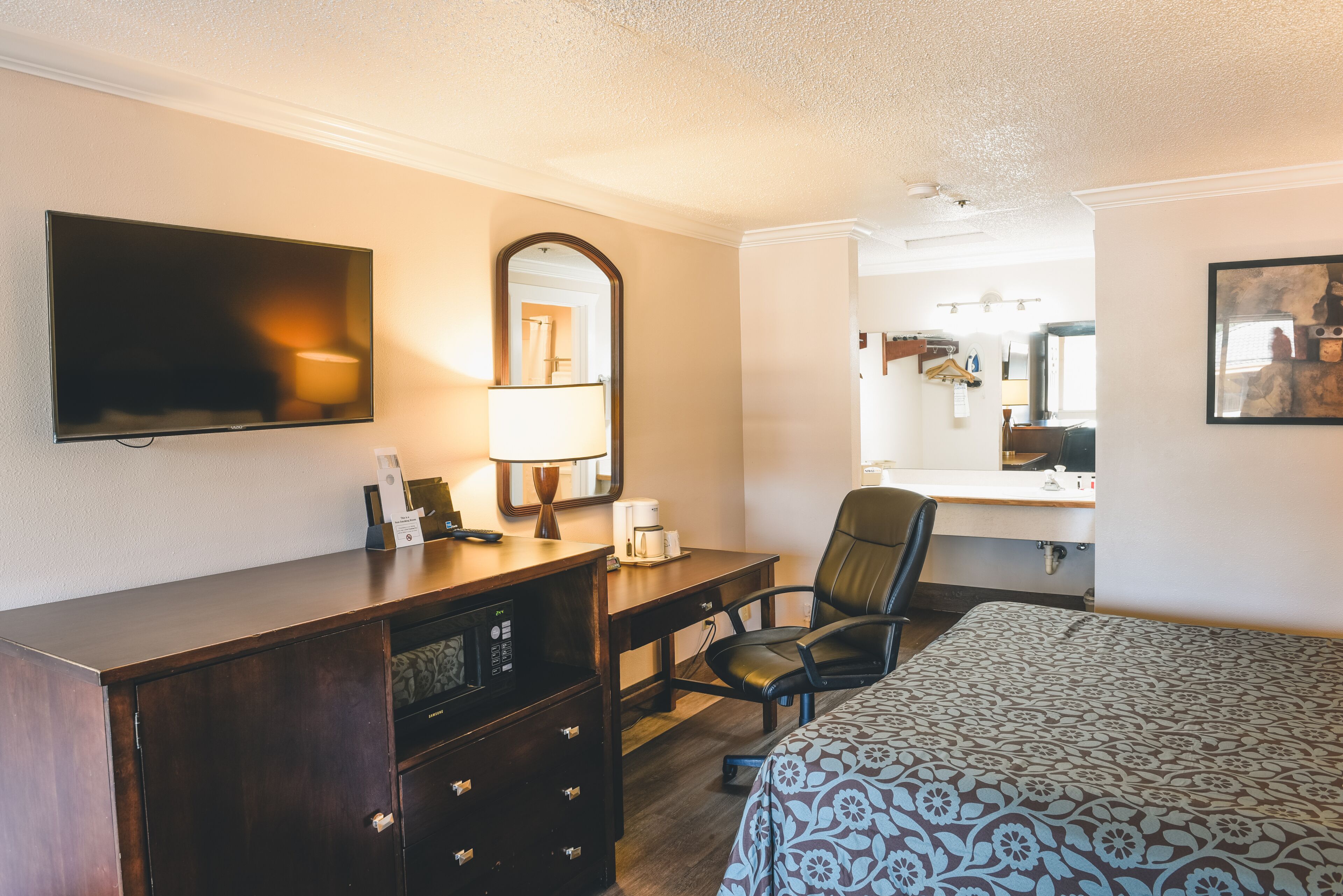 A hotel room with a bed, a desk, a chair, a TV, a mirror, and a dresser.