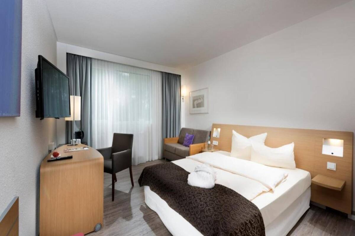 A hotel room with a bed, a desk, a chair, a TV, and a window with curtains.