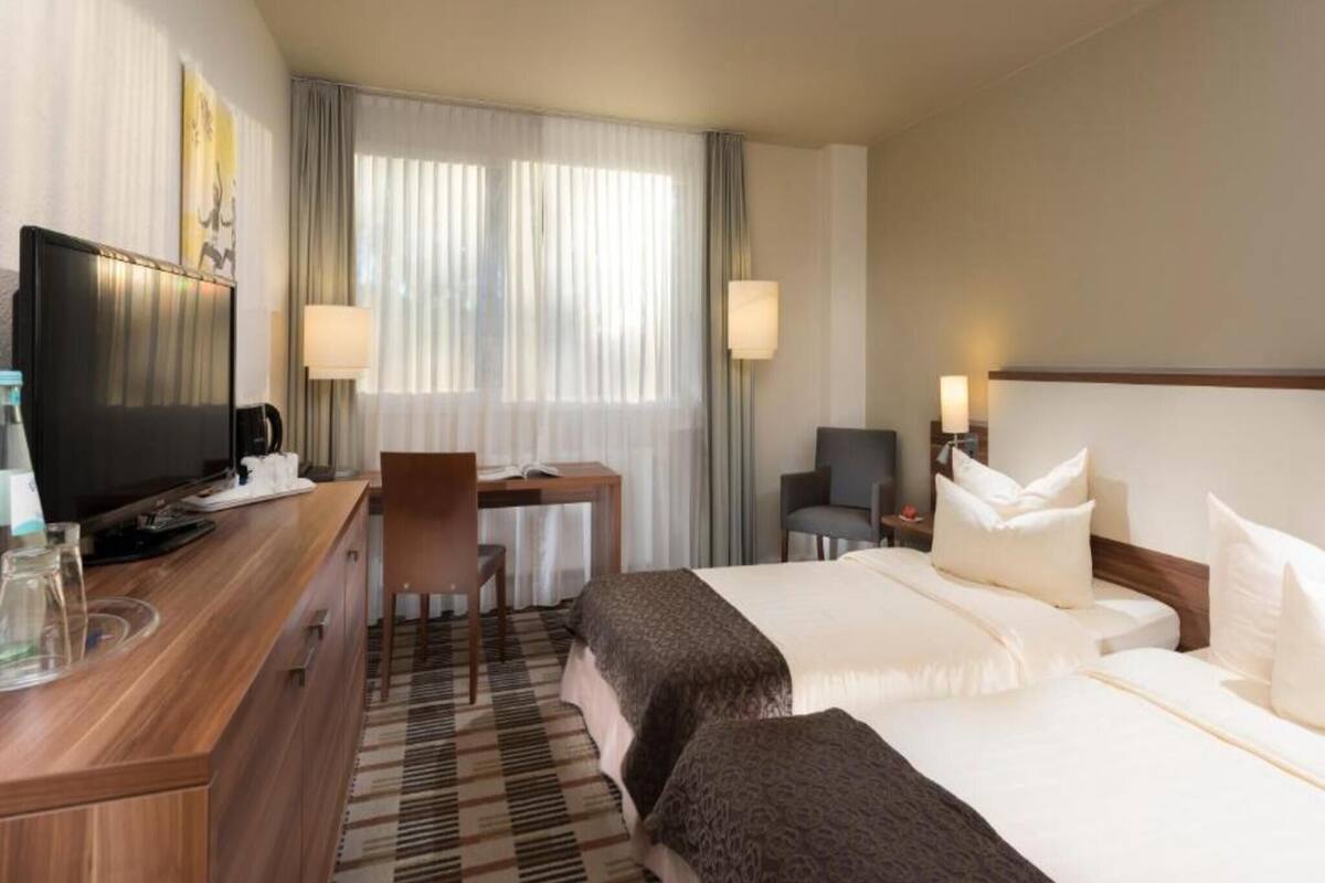 A hotel room with a bed, a desk with a chair, a television, and a window with curtains.