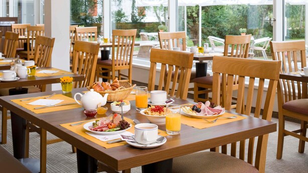 A breakfast setting with wooden tables, chairs, and a view of trees through large windows. - Sunday Hotel Saarbrücken Süd (Saarbrücken)
