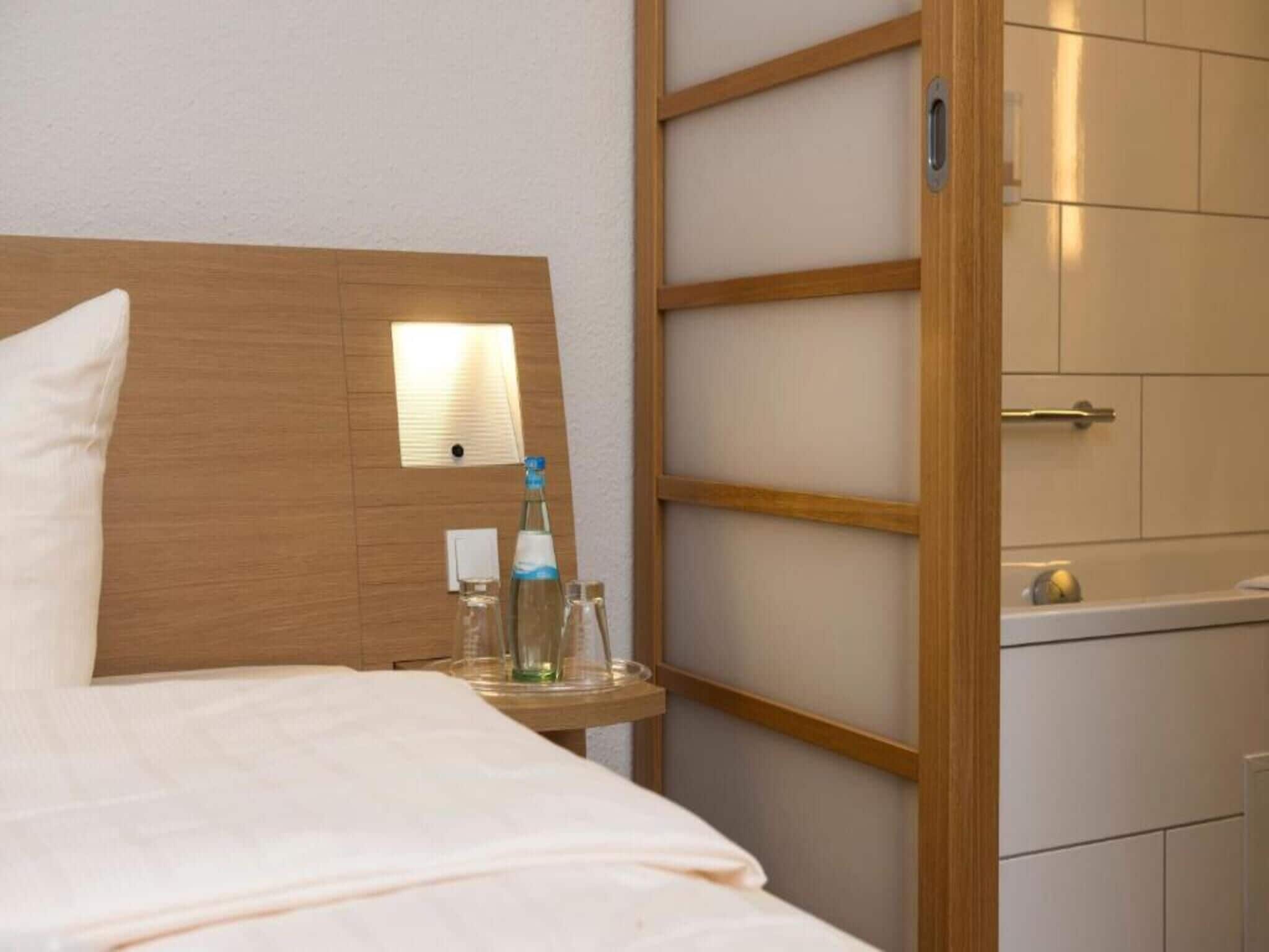 Hotellihuone, jossa on puinen sängynpääty, yöpöytä, jossa on pullo ja laseja, sekä pieni peili.