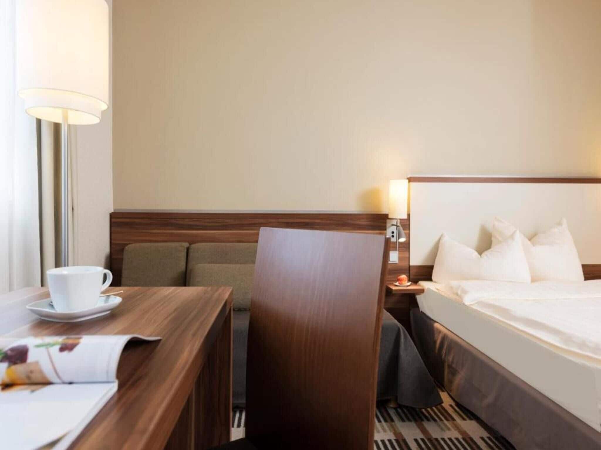 Ein Hotelzimmer mit Bett, Schreibtisch, Tasse und Untertasse, Stuhl und Lampe.