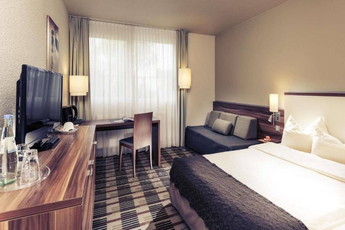 A hotel room with a bed, a desk with a TV, a chair, and a window with curtains.