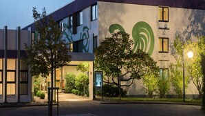 A modern building with a large green logo, a parking lot, and trees. - Sunday Hotel Saarbrücken Süd (Saarbrücken)