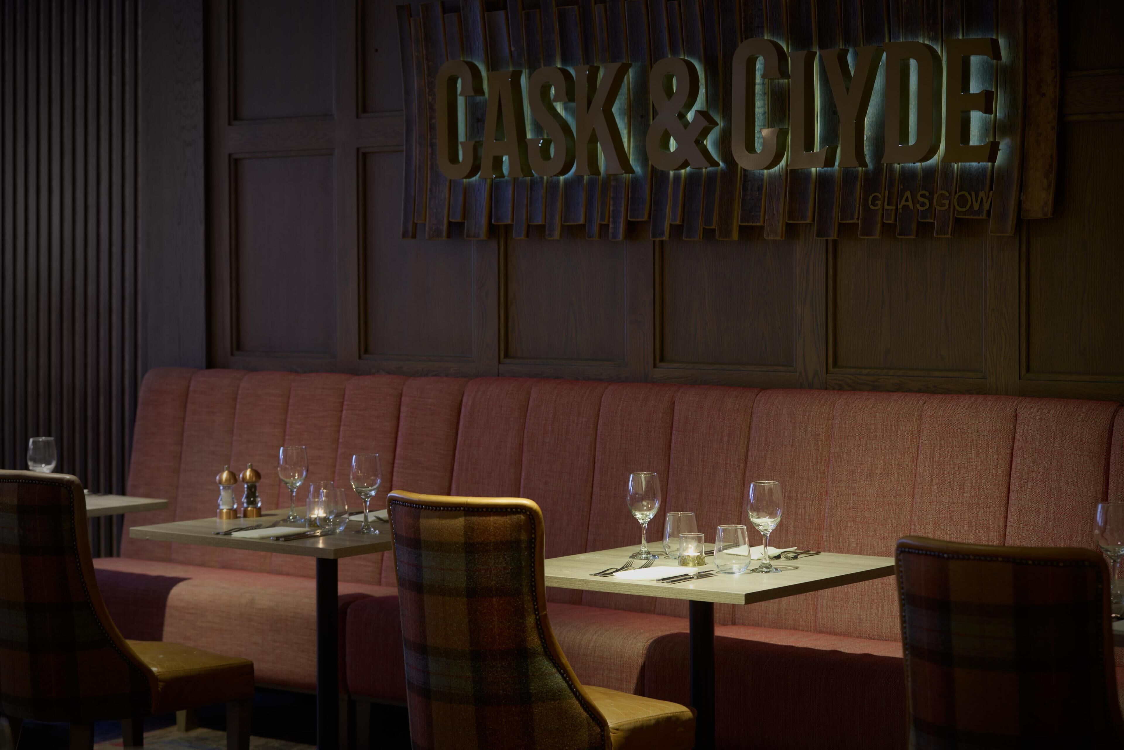 A restaurant interior with a red upholstered booth seating area, a wall-mounted sign reading 'CASK&CLYDE,' and tables set with glasses and cutlery.