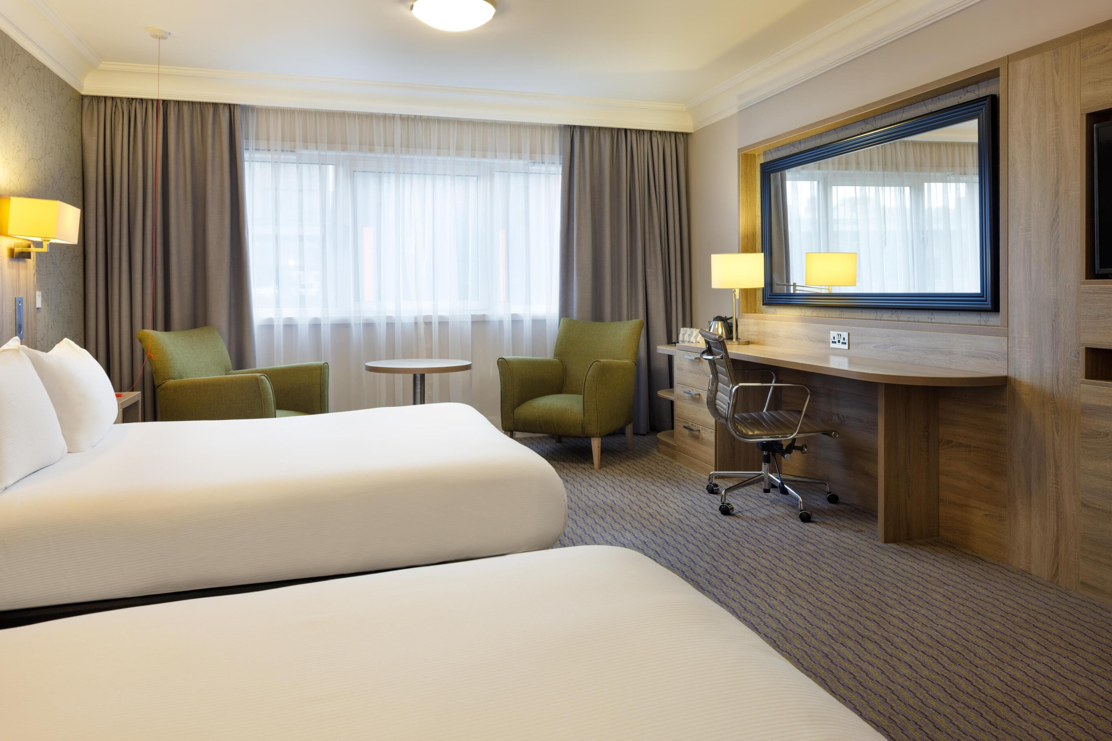 A hotel room with two beds, a desk, a chair, a small table, a lamp, and a large mirror.